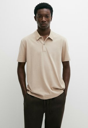 Jeune homme aux cheveux courts et bouclés porte un polo beige à manches courtes et un pantalon foncé, debout les mains dans les poches sur un fond uni.