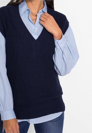 Persona che indossa un gilet smanicato lavorato a maglia blu navy sopra una camicia celeste con maniche lunghe a sbuffo e jeans blu.