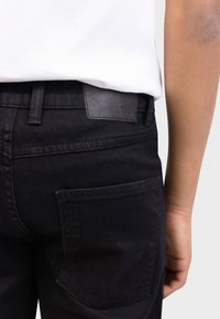 Μαύρο τζιν με ομαλή υφή, που διαθέτει πίσω τσέπη και δερμάτινη ετικέτα στη μέση. Φορεμένο με λευκό πουκάμισο.