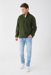 Grøn fleece pullover med høj krave, halv lynlås foran og en brystlomme. Kombineret med lyseblå jeans og hvide sneakers.