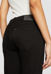 Svarta jeans med en figurnära design, slät textur och sydda detaljer. Midjebandet har en svart etikett och en guld logotypaccent.