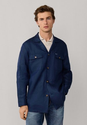 Young man wearing a navy blue button-up jacket over a light beige polo shirt and blue jeans, standing against a plain gray background.