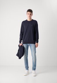 Tamsiai mėlyna apvalios iškirptės megztinis, šviesiai mėlynos džinsai, laiko tamsiai mėlyną striukę ir dėvi baltus sportbačius. Pagaminta iš medvilnės, gumuoti rankogaliai ir apykaklė.