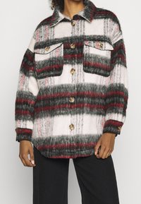 Rutigt skjortjacka med lång ärm i vitt, svart och rött, med en mjuk, luddfäst yta, två bröstfickor och stora beige knappar.