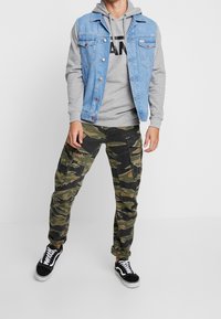 Gilet en denim bleu clair, sweat à capuche gris avec logo noir, pantalon cargo camouflage, et baskets noires avec semelles blanches. Style streetwear décontracté.