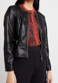 Veste noire en similicuir avec fermeture éclair devant et poches latérales, associée à un haut rouge à manches longues imprimé serpent. Coupe ajustée avec une texture lisse.