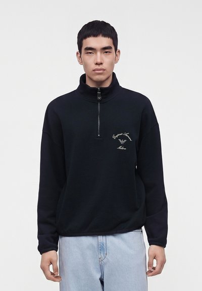 Jeune homme portant un sweat-shirt noir à fermeture quart de zip avec un texte de logo blanc sur la poitrine et un jean bleu clair, debout devant un fond uni.