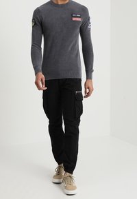 Grauer, gerippter Langarm-Pullover mit Aufnähern, kombiniert mit schwarzen Cargohosen mit Taschen und beigen Sneakern mit weißen Sohlen.