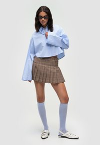 Camicetta croppata azzurro chiaro, gonna a pieghe scozzese marrone, calze blu chiaro fino al ginocchio e sandali bianchi con cinturini. Presenta maniche oversize.