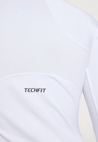 Bijela dugih rukava atletska majica s mrežastim panelom na gornjem dijelu leđa i crnim "TECHFIT" logotipom na donjem dijelu. Glatka tekstura.