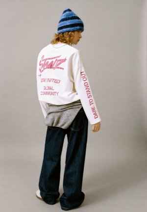 Witte sweatshirt met gedurfde roze tekst, grijs gelaagd kledingstuk, donkere spijkerbroek en blauwe gestreepte gebreide muts. Casual, ontspannen pasvorm.