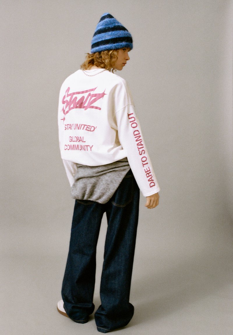 Felpa bianca con testo audace rosa, indumento grigio a strati, jeans scuri in denim e berretto a maglia a righe blu. Stile casual e taglio comodo.