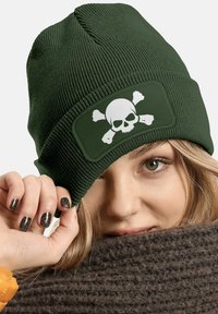 Green knitted beanie with a flat crown, ribbed texture, and a rectangular patch featuring a white skull and crossbones design.