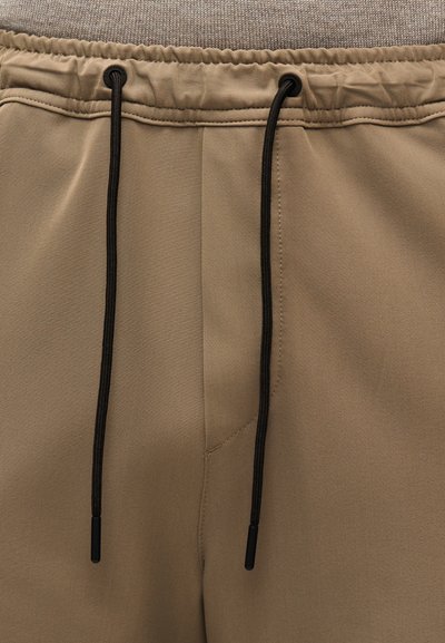 Gros plan sur la ceinture d'un pantalon kaki avec un haut élastique et des cordons noirs pendants.