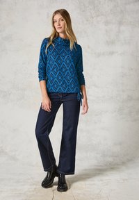 Blauwe coltrui met een ruitpatroon, gecombineerd met donkere denim flared jeans en zwarte enkellaarsjes, staand tegen een gestructureerde achtergrond.