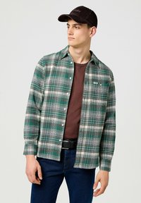 Grün-weiß kariertes Flanellhemd mit Knopfverschluss, über einem braunen T-Shirt getragen, kombiniert mit dunkelblauen Jeans und einer braunen Kappe.