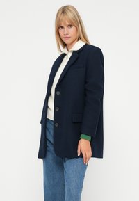 Donna bionda che indossa un cappotto blu navy con bottoni sopra un maglione bianco con zip e jeans blu, in piedi su uno sfondo bianco semplice.