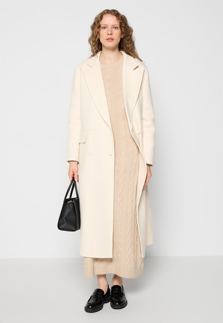 Langer cremefarbener Wollmantel über einem beigen Kabelstrickkleid, kombiniert mit einer schwarzen Umhängetasche und schwarzen Loafers. Glatte Texturen und klassische Linien.