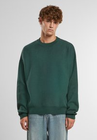 Dunkelgrüner Strickpullover mit Rundhalsausschnitt, überschnittenen Schultern und gerippten Bündchen und Saum. Kombiniert mit hellblauen Jeans.