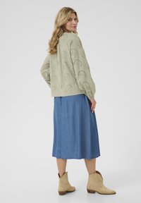 Hellgrüne, gemusterte Bluse mit gesticktem Design, kombiniert mit einem Jeansrock und beigen Ankle Boots, von hinten betrachtet.