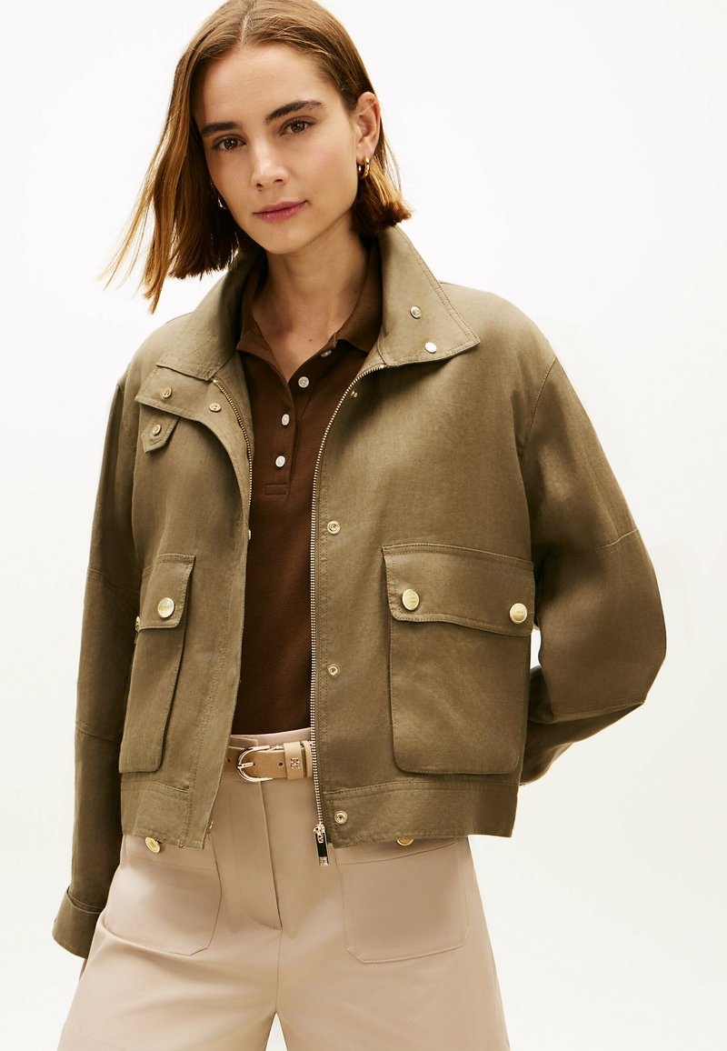 Woman with shoulder-length hair wearing an olive-green jacket with large flap pockets, zipped open over a brown polo shirt and beige pants.