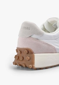 Bílé tenisky s růžovým a šedým semišovým podpatkem, texturovanou hnědou gumovou podrážkou a hladkým syntetickým svrškem. Obsahují hexagonální hroty pro zajištění trakce.