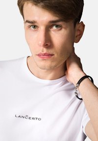 Biała koszulka z logo "LANČERTO" na piersi. Osoba ma na sobie czarny pleciony bransoletkę z metalowymi akcentami.