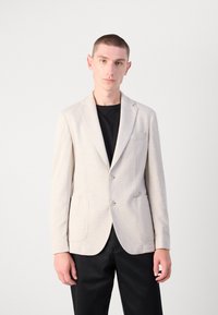 Blazer beige chiaro con un sottile motivo a scacchi, due tasche frontali e due bottoni, indossato su una camicia nera e abbinato a pantaloni scuri.