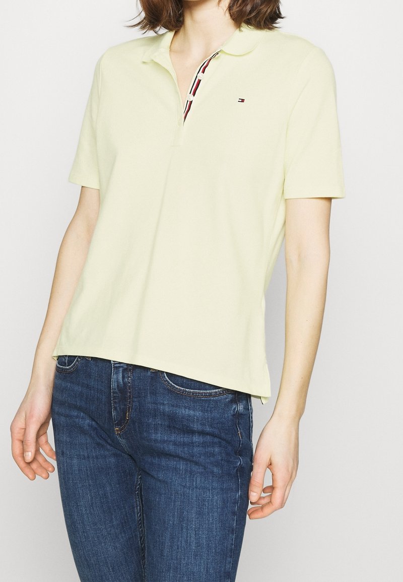 Femme portant un polo à manches courtes jaune pâle avec un petit logo et un jean bleu, sur un fond blanc uni.