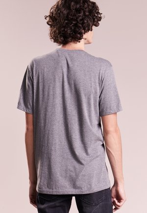 Personne aux cheveux bouclés portant un t-shirt gris uni à manches courtes, de dos, debout devant un fond beige neutre.