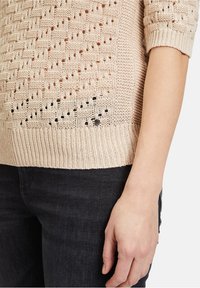 Gestrickter hellbeiger Pullover mit offenen Strickmustern, gerippten Bündchen und Saum. Kombiniert mit dunklen Jeans für einen Kontrast.