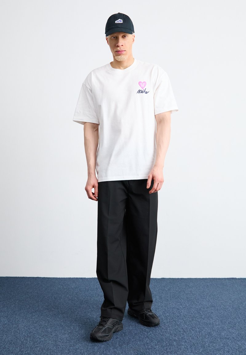 Camiseta blanca de algodón con un gráfico de un corazón y el texto "Nike" en rosa, combinada con pantalones holgados negros y zapatillas negras. Gorra negra.