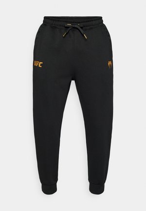 Pantaloni della tuta neri con gambe affusolate, vita elasticizzata e coulisse. Presentano il logo UFC arancione sulla coscia sinistra e un logo sulla destra.
