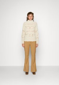 Romantisch gebreid sweater met opengewerkte patroon, ribgebreide manchetten, gecombineerd met beige flared broek en bruine haklaarzen, staande tegen een effen achtergrond.