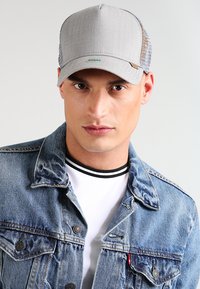 Gorra gris con un frente de tela texturizada y parte trasera de malla; presenta un logo verde y un visera curvada. Llevada con una chaqueta de mezclilla y una camisa blanca.
