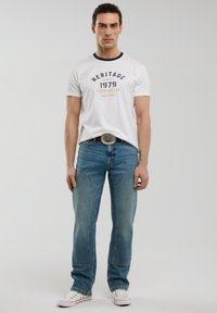 Balta crewneck marškinėliai su mėlyna apdaila ir spausdintu tekstu, derinami su šviesiai mėlynomis džinsomis, turinčiomis mažą išblukimą ir siuvimo detales, baltais sportbačiais.