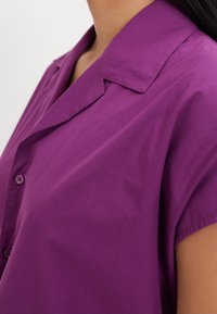 Maglietta viola a maniche corte con colletto classico e chiusura frontale a bottoni. Realizzata in tessuto morbido con una texture liscia e un fit comodo.