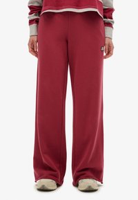 Person, der lockere mahagonifarbene Sweatpants, beigefarbene Sneaker und ein langärmliges Oberteil in Marmor- und Grautönen trägt, steht mit entspannten Händen an den Seiten.