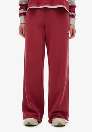 Person wearing loose maroon sweatpants, beige sneakers, and a maroon and gray long-sleeve top, standing with hands relaxed by sides.