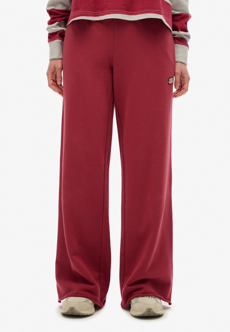 Person, der lockere mahagonifarbene Sweatpants, beigefarbene Sneaker und ein langärmliges Oberteil in Marmor- und Grautönen trägt, steht mit entspannten Händen an den Seiten.