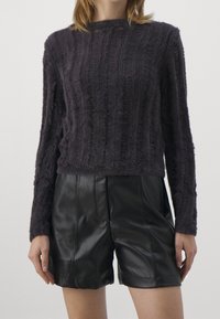 Person, der einen dunklen, flauschigen Strickpullover und hoch taillierte schwarze Leder-Shorts trägt, vor einem schlichten Hintergrund.