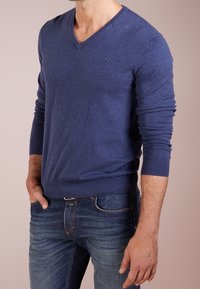 Pull bleu marine à col en V en tissu tricoté doux avec manches longues, poignets et ourlet côtelés, associé à un jean en denim bleu.