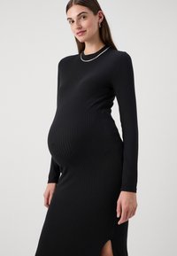 Μαύρο ριγέ φορέμα εγκυμοσύνης με μακριά μανίκια, στρογγυλή λαιμόκοψη και πλευρικό άνοιγμα. Το ύφασμα φαίνεται ελαστικό και απαλό, εφαρμόζοντας στενά στο σώμα.