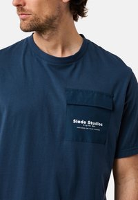 T-paita tummansinisestä puuvillasta, jossa on etupuolella läpällä varustettu tasku, kohokuvioitu teksti ja pyöreä kaula-aukko. Sileä pinta.