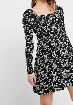 Woman wearing a black long-sleeve dress with white daisy floral print, smocked bodice, and flared skirt.