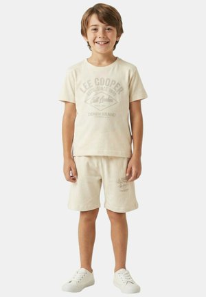 Garçon souriant debout, portant un t-shirt beige Lee Cooper et un short assorti avec des baskets blanches, sur un fond clair uni.
