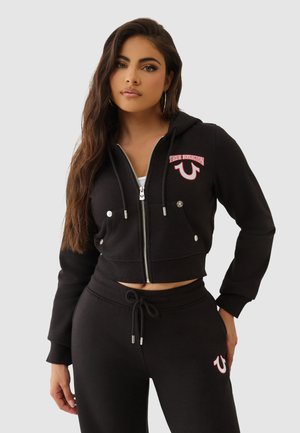 Schwarze, kurze Zip-Hoodie mit silberner Hardware, ausgestattet mit einem roten gestickten Logo und zwei vorderen Taschen, kombiniert mit passenden Jogginghosen.