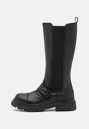 Botte haute en cuir noir avec semelle épaisse, panneau latéral élastique, et bride décorative avec boucle métallique autour de la cheville.