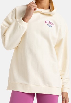 Persoon draagt een crèmekleurige oversized hoodie met roze "ROXY"-tekst en logo op de borst, gecombineerd met felpaarse leggings.