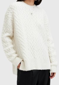 Personne portant un pull blanc en maille torsadée avec poignets et ourlet côtelés, associé à un pantalon noir, les mains partiellement dans les poches.